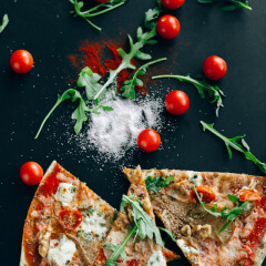 Porciones de pizza con queso y rúcula sobre fondo negro, rodeadas de tomates cherry, hojas verdes, sal y pimentón en polvo.