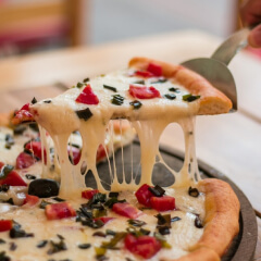 Rebanada de pizza con queso fundido estirándose, aceitunas negras y trozos de tomate, siendo servida sobre bandeja de madera.
