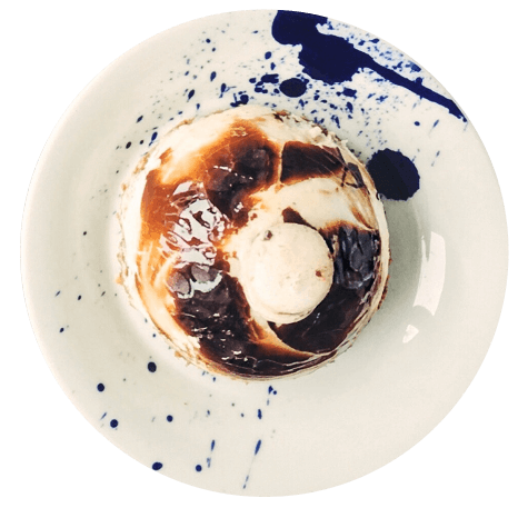 Postre redondo con base cremosa y cobertura de chocolate marmoleado, servido en plato blanco con salpicaduras decorativas de color azul oscuro.