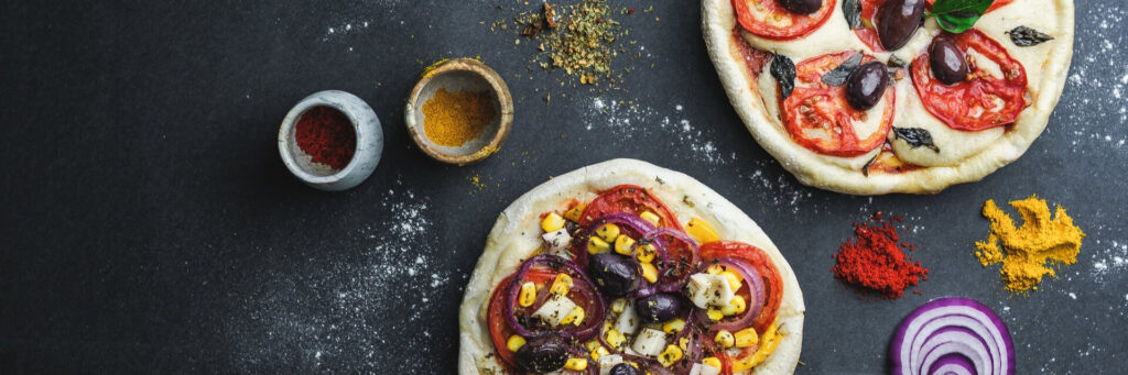 Dos mini pizzas sobre fondo oscuro, una con tomate, aceitunas y albahaca, y otra con maíz, cebolla y queso, rodeadas de especias y vegetales.