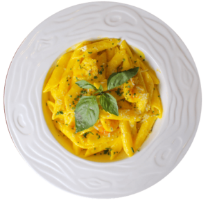 Plato hondo blanco con pasta penne en salsa amarilla cremosa, decorada con camarones, perejil picado y hojas de albahaca fresca.
